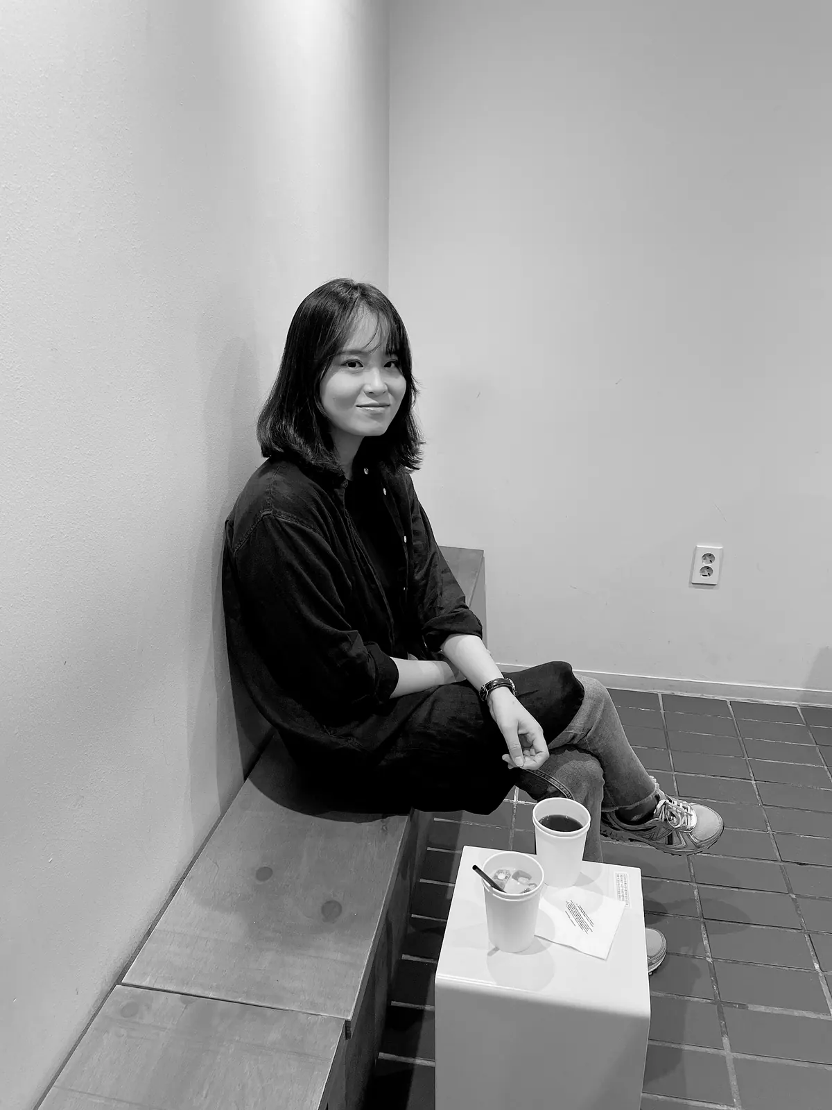 하얀 벽 앞 나무 벤치에 앉아 카메라를 향해 미소 짓는 김진